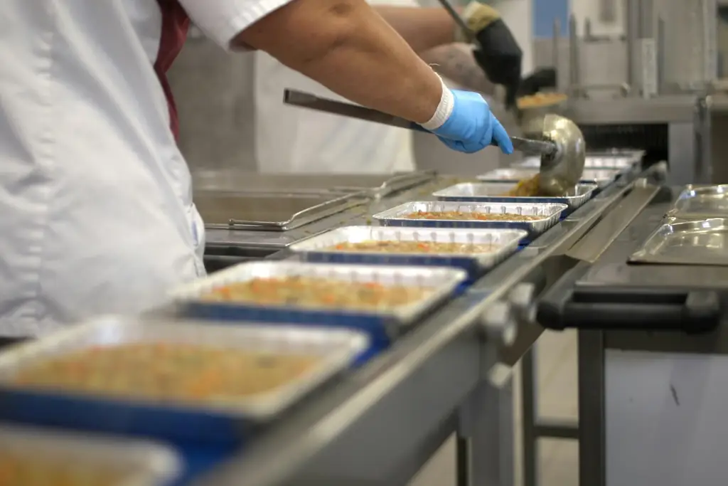 Una persona reparte comida en bandejas sobre una línea de producción.